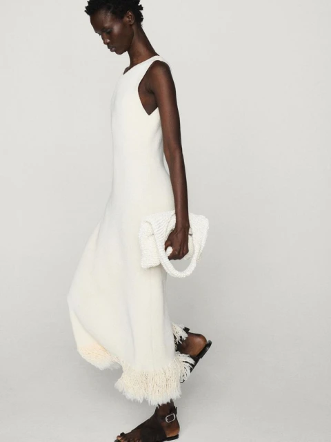 Elegant African American woman in a cream-colored dress and sandals, holding a beaded purse against a neutral studio background.