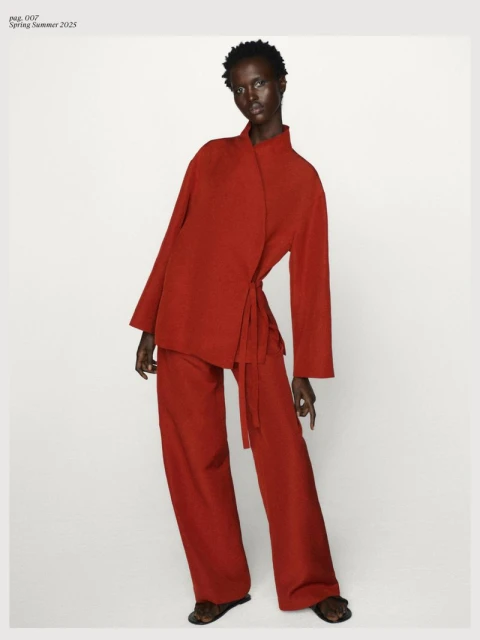 Fashion editorial shot of an African female model in a red wrap jacket and wide leg pants, posing against a plain background, styled professionally.
