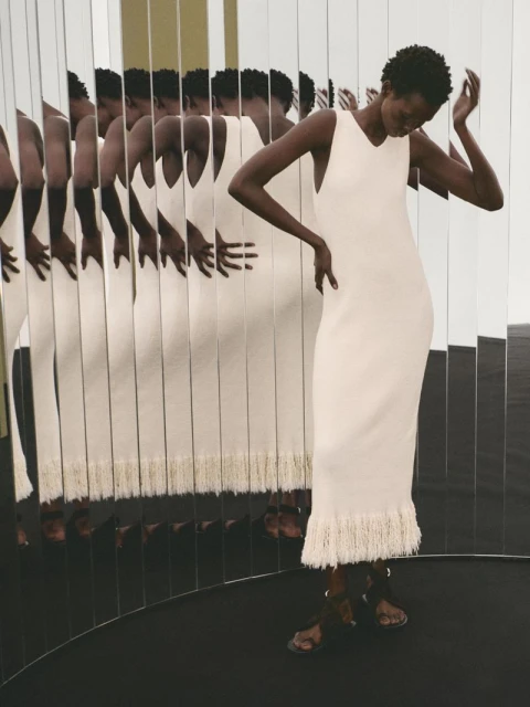 Elegant woman in a white dress poses in a mirrored room, creating multiple reflections. The dress has fringe detail at the hem.