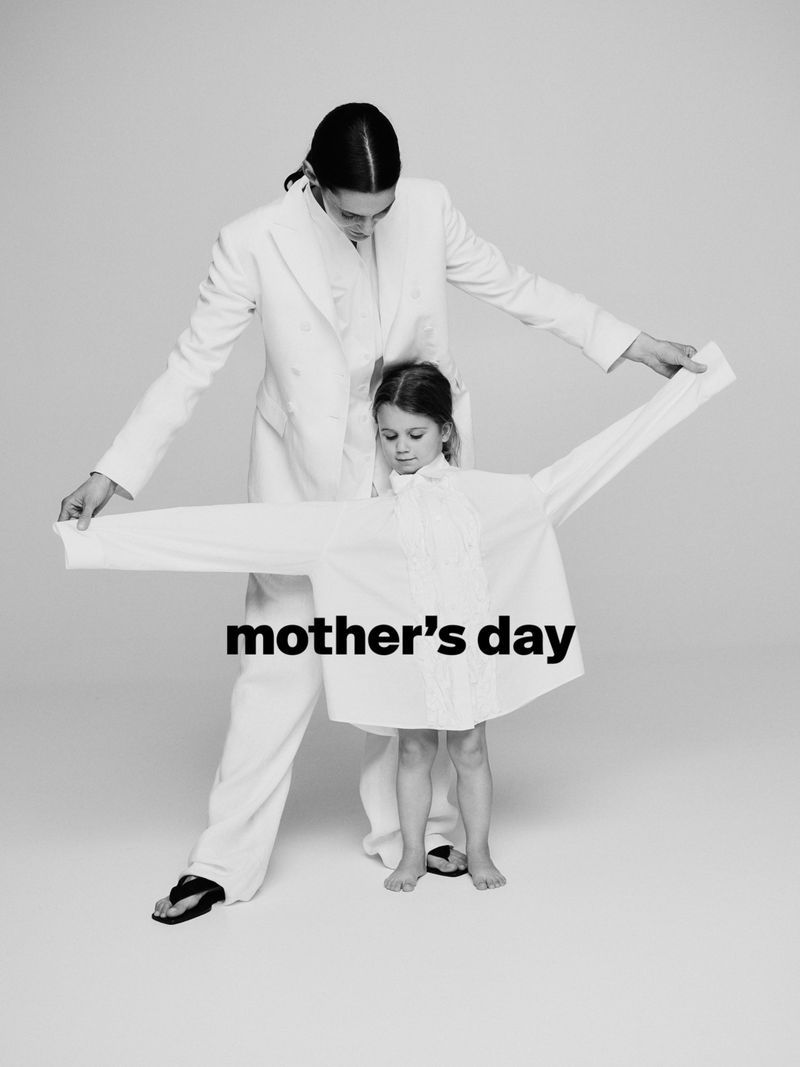 A woman and a child stand indoors against a plain backdrop, both wearing white shirts, in a minimalist, black-and-white fashion shot.
