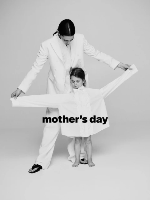 A woman and a child stand indoors against a plain backdrop, both wearing white shirts, in a minimalist, black-and-white fashion shot.