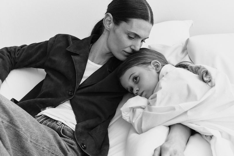 Woman in dark jacket leaning over a child resting on white pillows in a black and white portrait