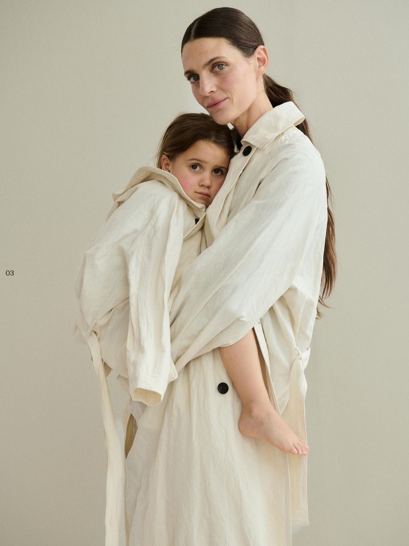 Mother and child in matching beige linen trench coats in a studio setting. The child is held closely by the adult, both looking at the camera.