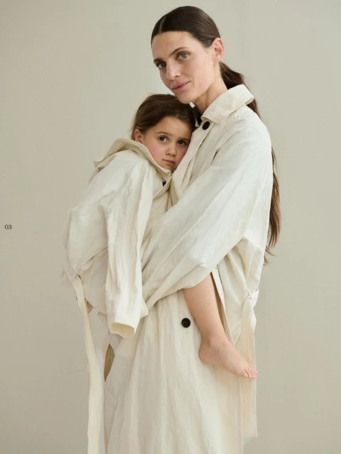 Mother and child in matching beige linen trench coats in a studio setting. The child is held closely by the adult, both looking at the camera.