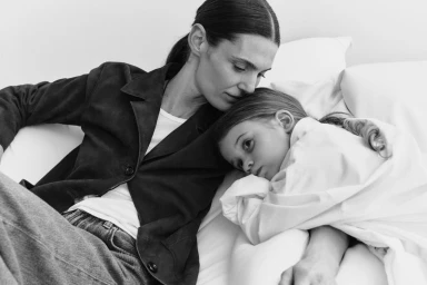 Woman in dark jacket leaning over a child resting on white pillows in a black and white portrait
