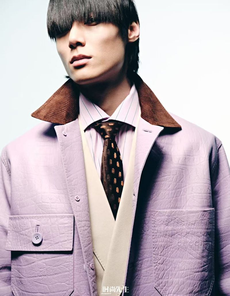 Close-up of an adult male model wearing a lilac jacket with a brown corduroy collar, a striped shirt, and a patterned tie in a studio.