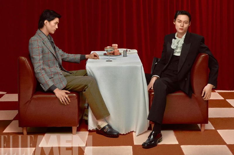 Two adult males seated at a table with drinks and desserts in a photo studio against a red curtain backdrop and checkered floor.