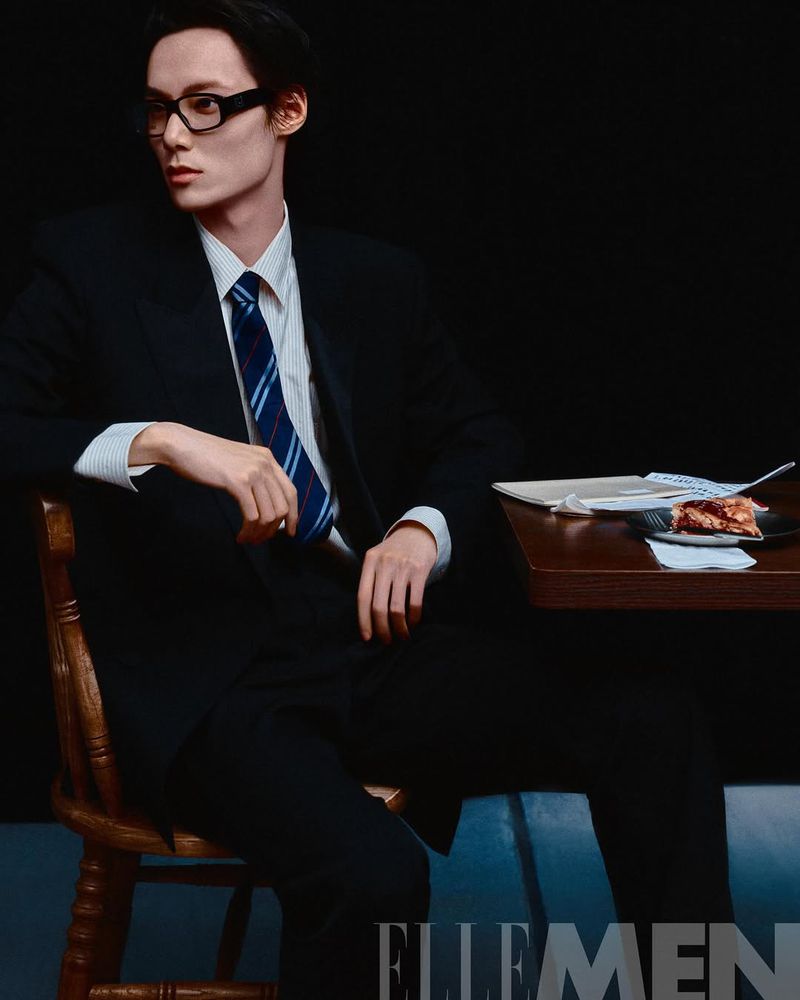 An adult male model in a black suit and rectangular glasses sitting at a table next to a plate of food, looking at the camera.
