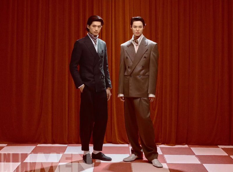 Two stylish men in suits pose in front of a red curtain on a checkered floor, showcasing a blend of classic tailoring and modern fashion.