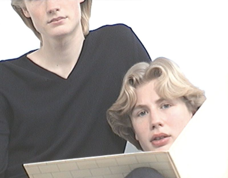 Two young adult males are captured indoors, one in a black sweater holding a tiled object, with a clean white background highlighting their features.