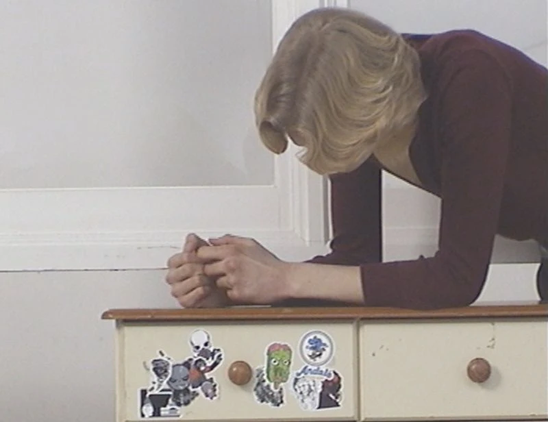 Blonde woman leans on a dresser with cartoon stickers near a window. She wears a maroon top in an indoor setting.