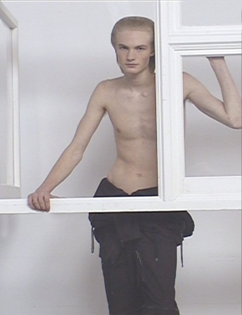 Nude young adult man in dark pants stands in a white frame indoors against a solid white background, portrait style. 
