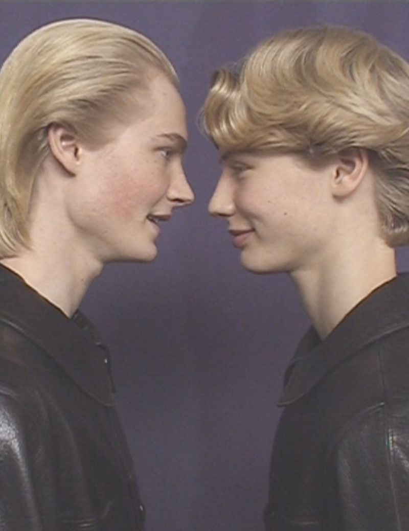 Close up of two fair-skinned young adult men looking at each other, wearing black jackets against a blue background in a studio.
