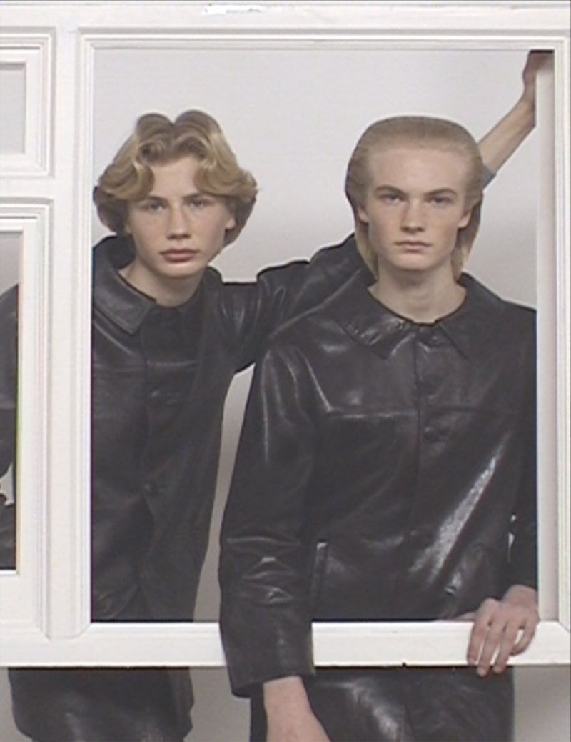 Two young men in matching black leather jackets pose within a white rectangular frame against a bright white studio backdrop.
