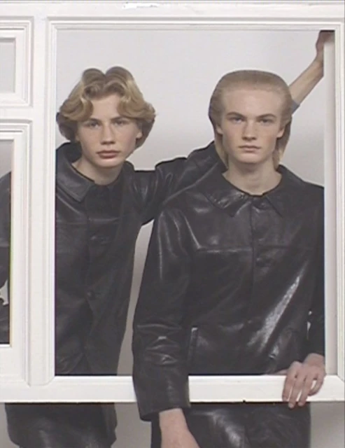Two young men in matching black leather jackets pose within a white rectangular frame against a bright white studio backdrop.