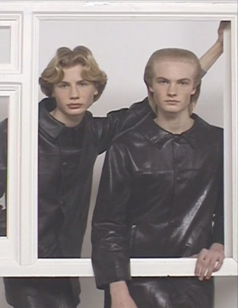 Two young men in matching black leather jackets pose within a white rectangular frame against a bright white studio backdrop.