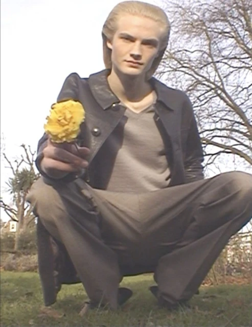 Young adult male with blond hair squats on green grass outdoors, offering a yellow flower toward the viewer in a medium shot with bare trees.