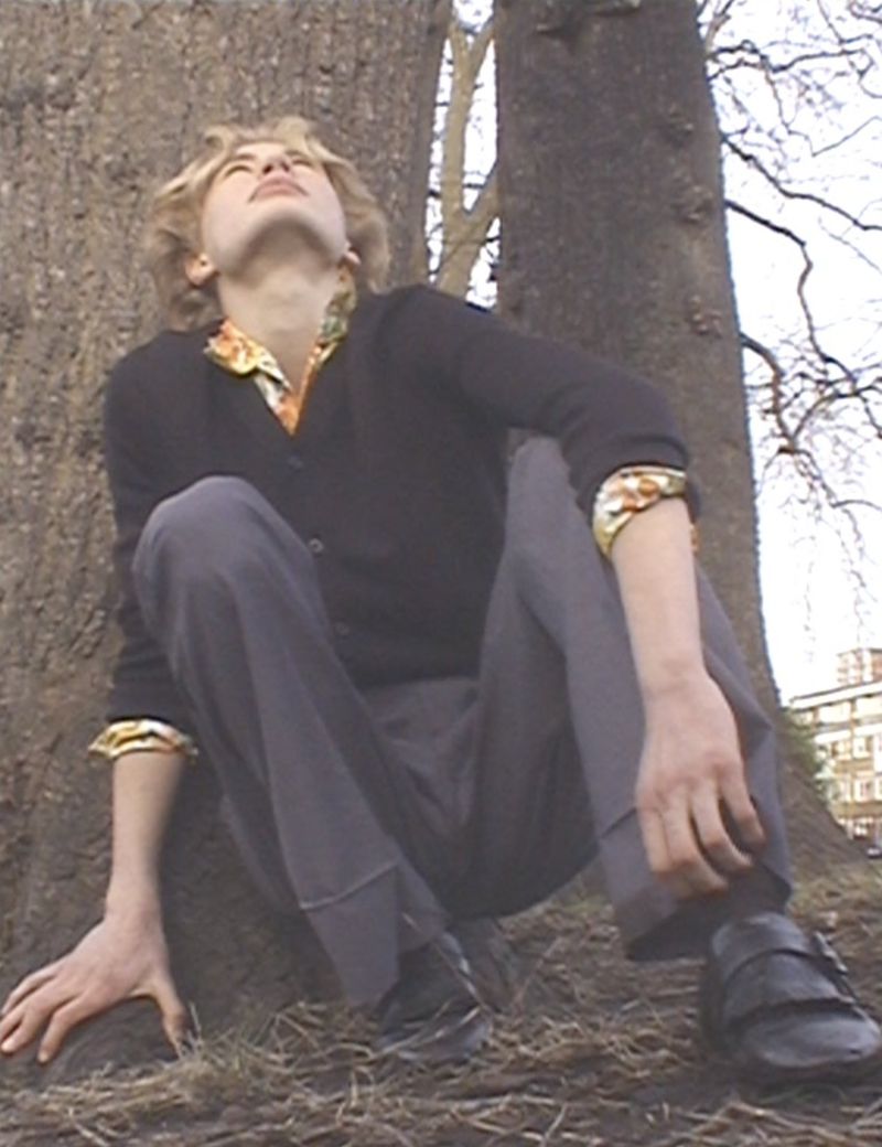 Young adult crouches near a tree on the ground. The individual wears casual attire including a sweater, trousers, and dress shoes.