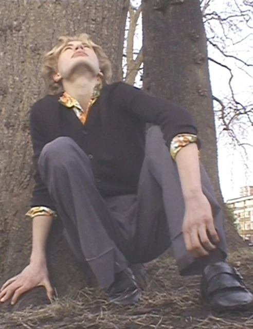 Young adult crouches near a tree on the ground. The individual wears casual attire including a sweater, trousers, and dress shoes.