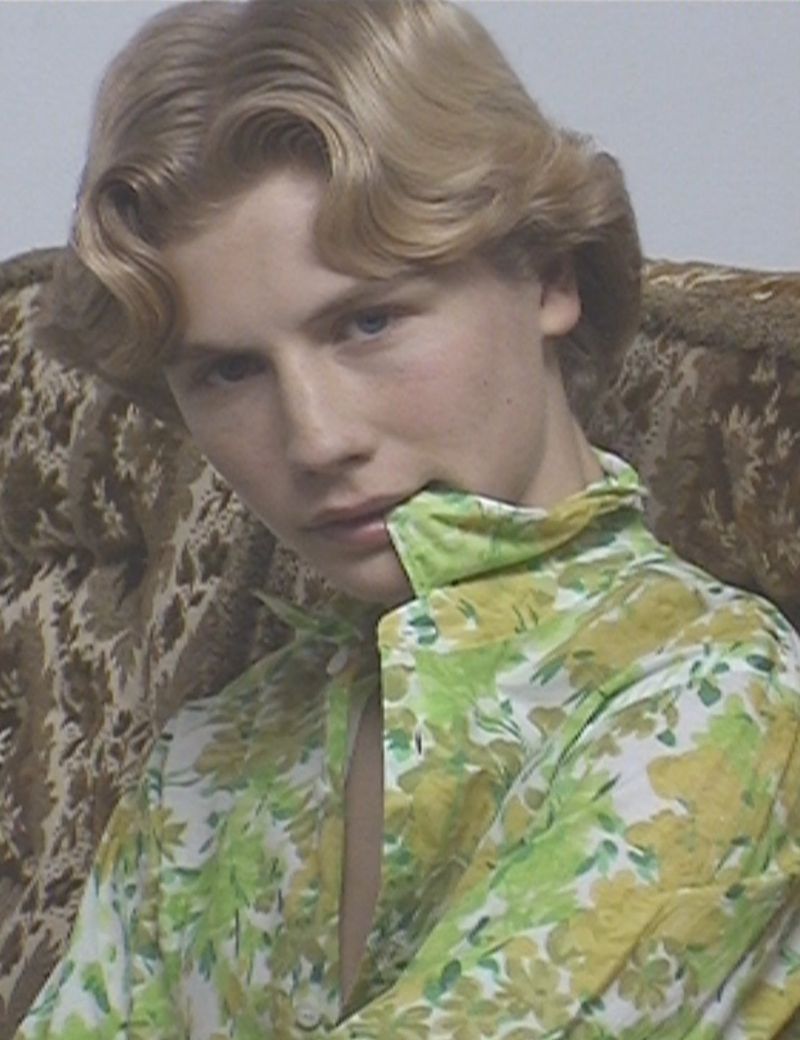 Close-up portrait of a young adult with blonde wavy hair wearing a green and yellow floral shirt looking at the camera indoors.