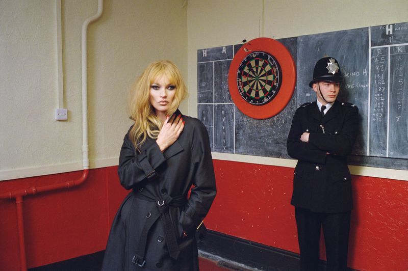 Woman with blonde hair in a black coat poses indoors with a British policeman standing against a chalkboard in the background.