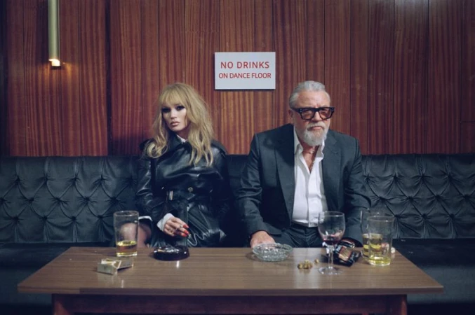 A woman and an older man seated indoors at a table with drinks, exuding a vintage aesthetic with wood paneling and soft lighting.