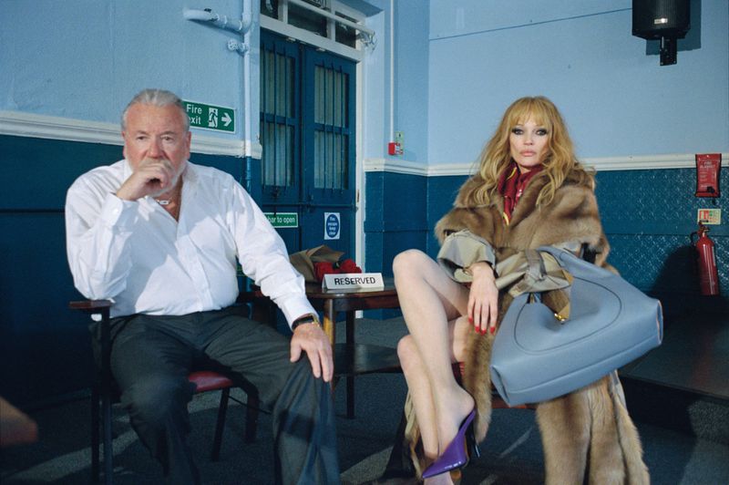 Two adults, a man and a woman, sit in chairs in an indoor setting with blue walls and dark blue door. The woman holds a large blue purse.