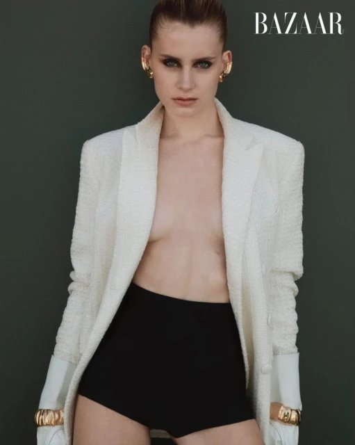 Fashion editorial shot of an adult female model with slicked-back hair wearing a white blazer and black shorts against a dark green backdrop.