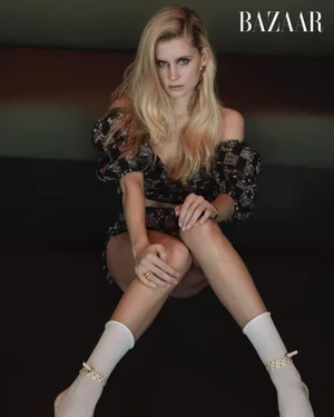 Blonde woman in dark patterned two piece outfit and white socks seated against a dark interior background