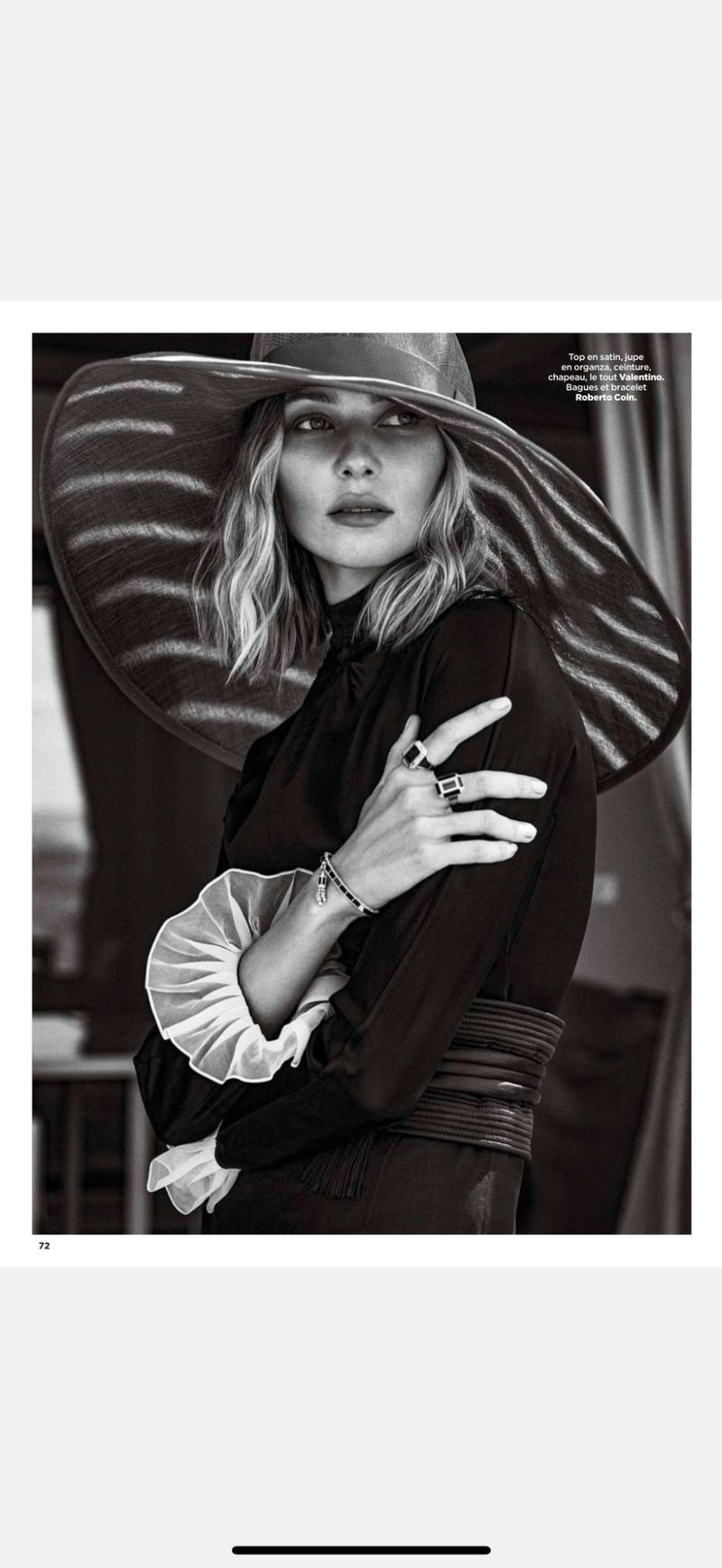 A woman with fair skin and wavy blonde hair wears a wide-brimmed sunhat and long-sleeved black dress with a ruffled collar.