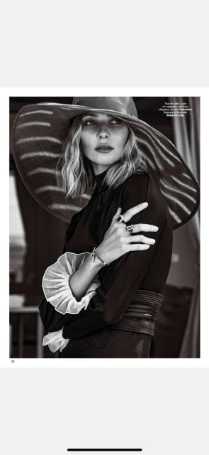 A woman with fair skin and wavy blonde hair wears a wide-brimmed sunhat and long-sleeved black dress with a ruffled collar.