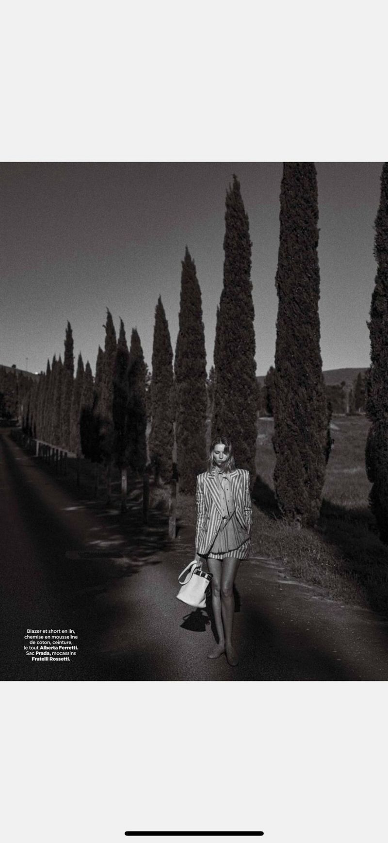 A woman walks along a tree-lined road in a black and white fashion shot, carrying a white purse, evoking a vintage or timeless feel.