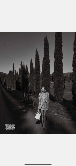 A woman walks along a tree-lined road in a black and white fashion shot, carrying a white purse, evoking a vintage or timeless feel.