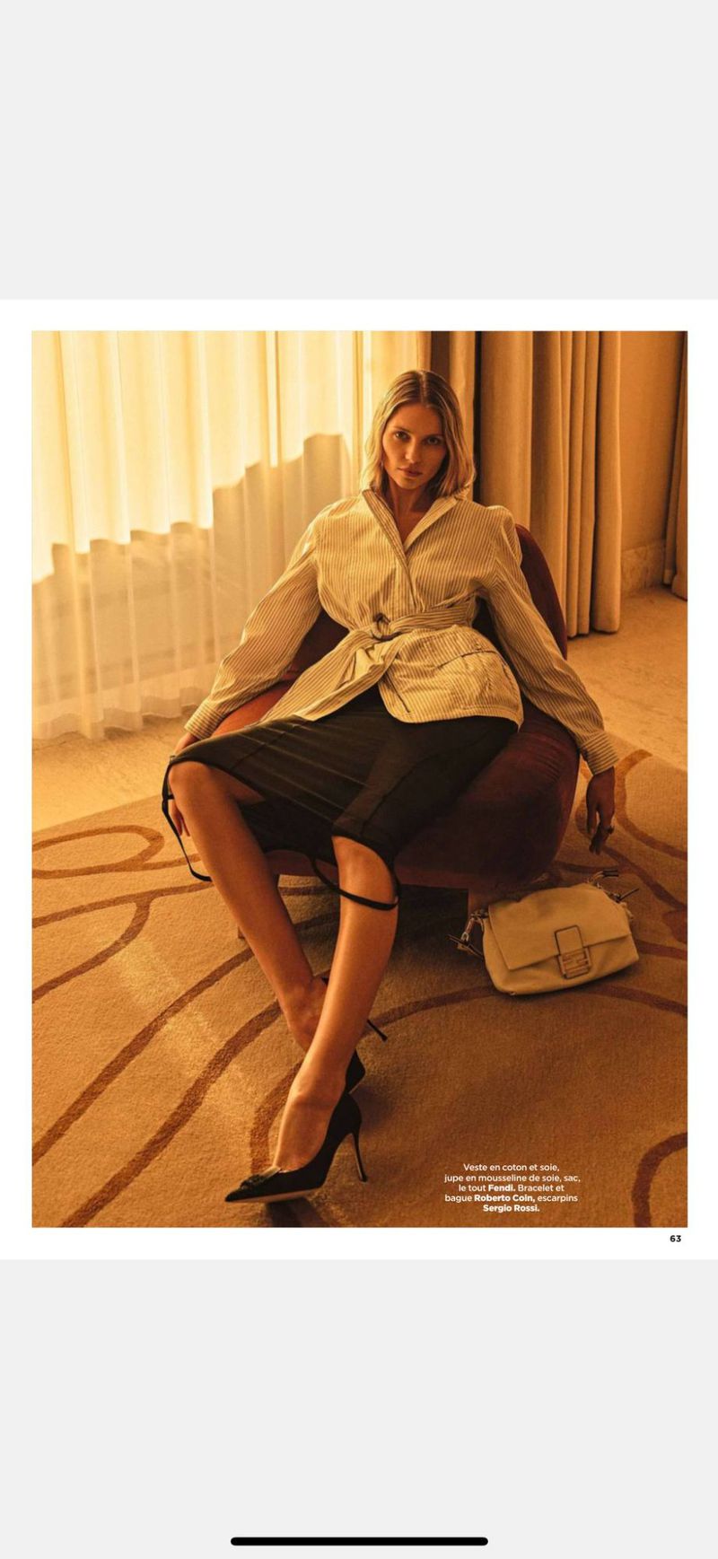 Fashion editorial shot of a blonde woman in a white jacket and black skirt, seated on a chair in a room with tan carpet.