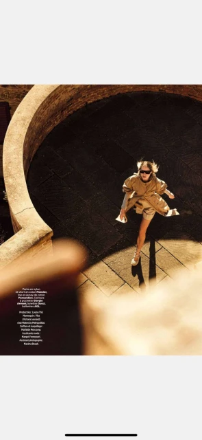 Woman in stylish tan outfit, sunglasses, and heels walking across shadowed cobblestones with curved stone railing on a sunny day.