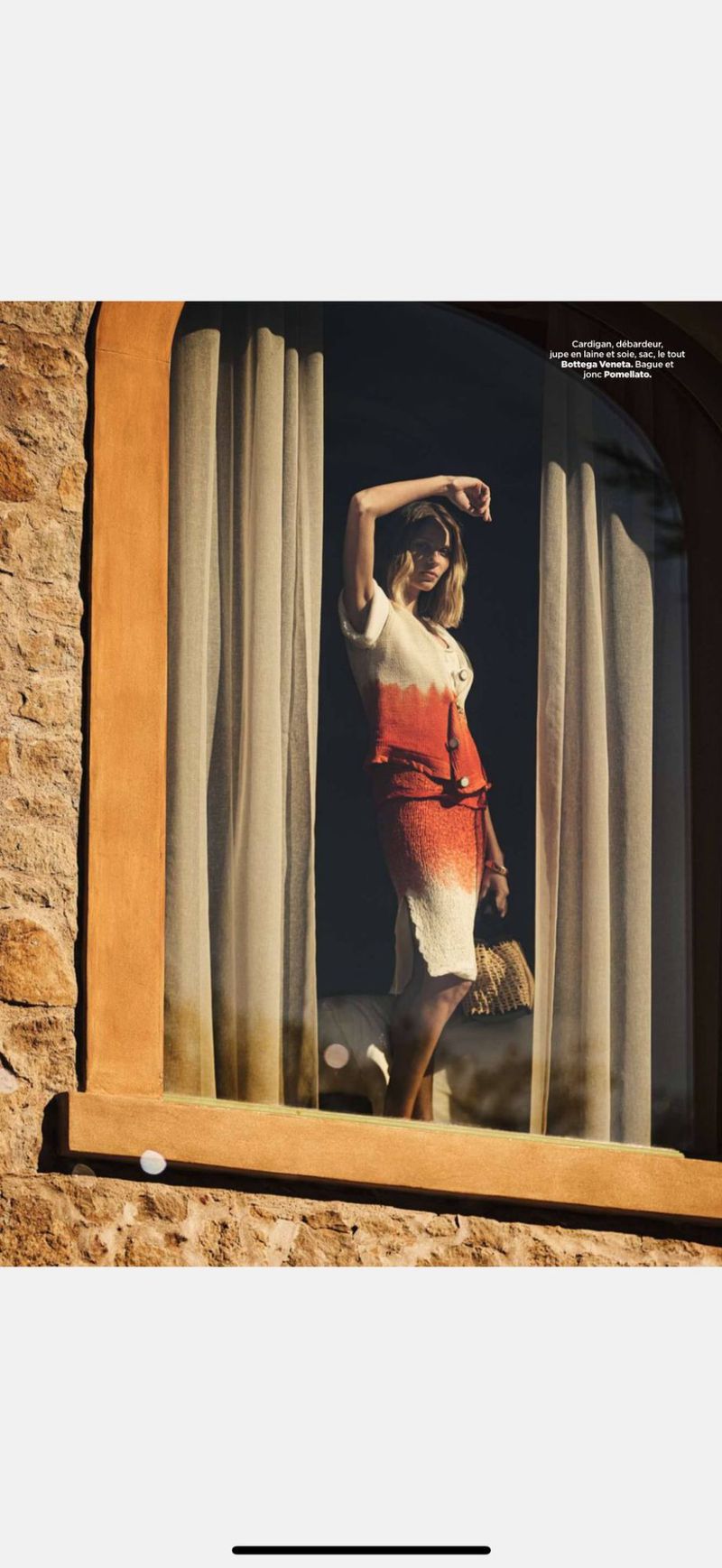 Woman in a vintage-style dress stands in a window, holding a straw purse, bathed in sunlight with stone walls in the background.