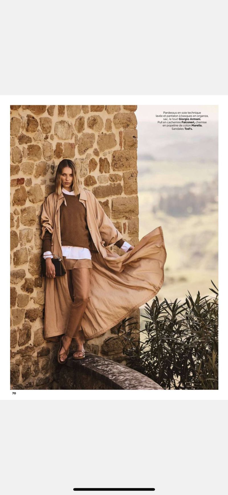 Fashion shot of a woman in earth-toned clothing with a flowing coat, posing against a stone wall in a blurred landscape setting.