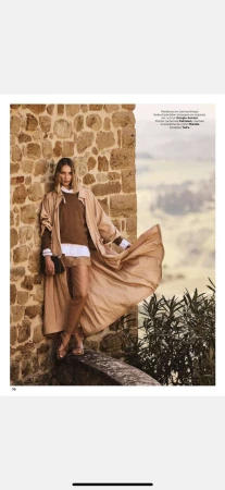 Fashion shot of a woman in earth-toned clothing with a flowing coat, posing against a stone wall in a blurred landscape setting.