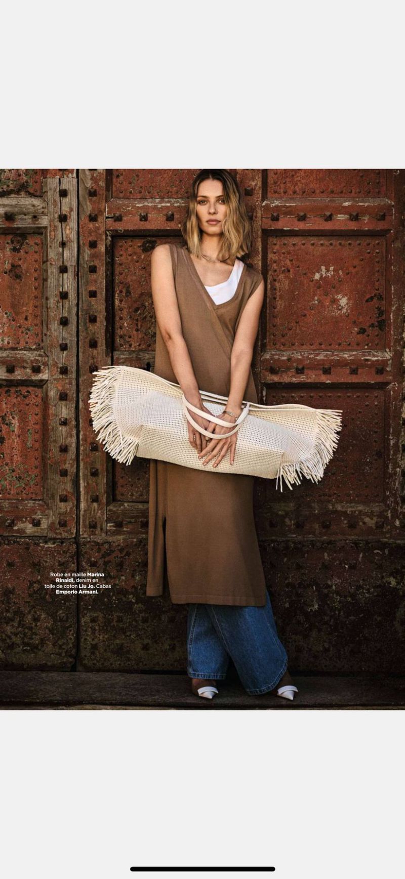 A woman in a brown sleeveless dress poses against a large rusty metal door while holding a fringed, woven tote bag in an editorial fashion image.