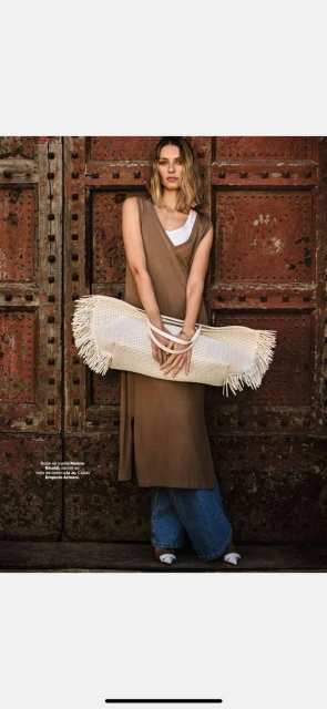A woman in a brown sleeveless dress poses against a large rusty metal door while holding a fringed, woven tote bag in an editorial fashion image.