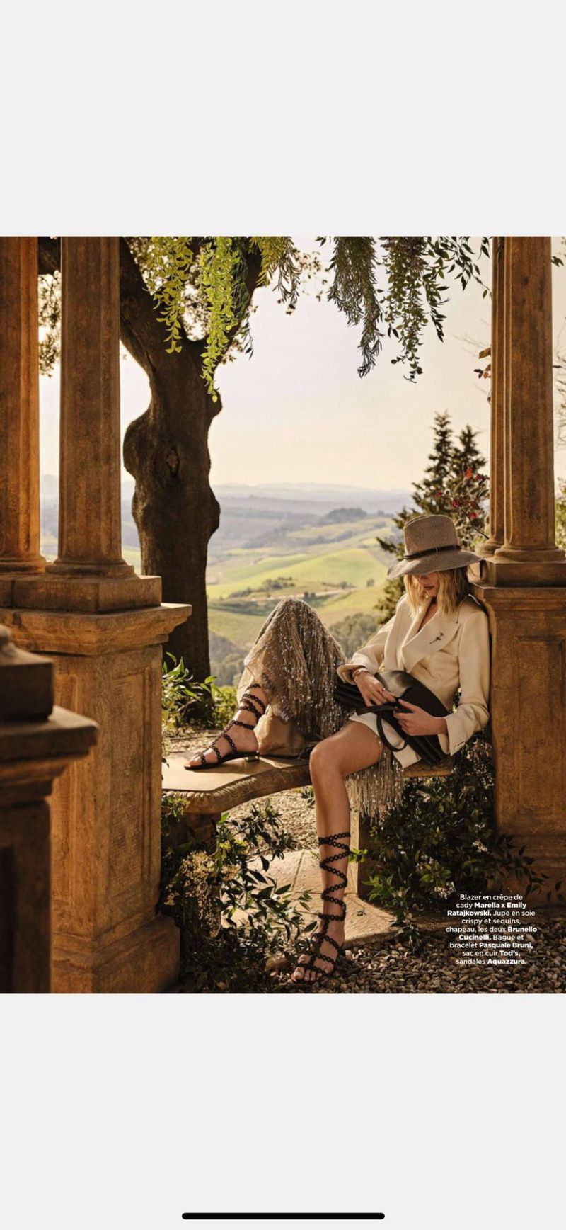Woman seated in a stone gazebo overlooking a green, hilly landscape, wearing a light-colored blazer, hat, and gladiator sandals.