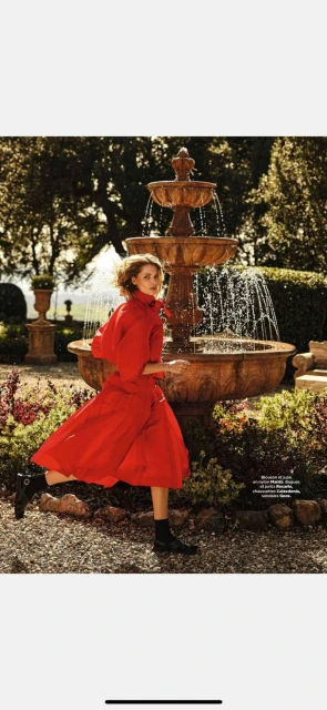 A fair-skinned young adult female model in motion poses in a red dress and black boots next to a stone fountain in a garden setting.