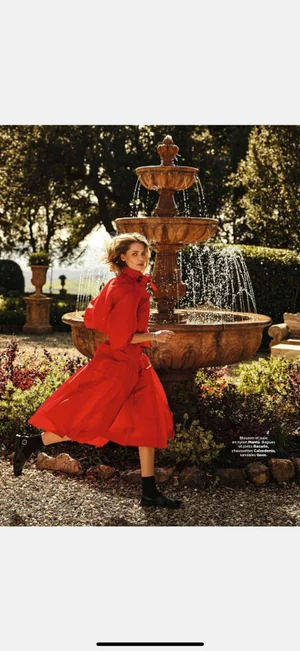 Model wearing a vibrant red dress running past an ornate outdoor stone water fountain in a lush garden