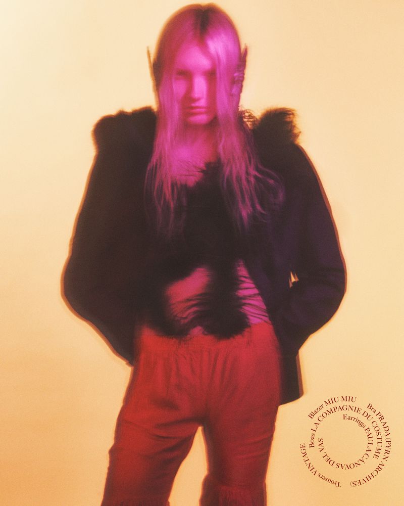 Portrait of a slender adult female with long blonde hair wearing a black jacket and red pants against a diffused orange background.