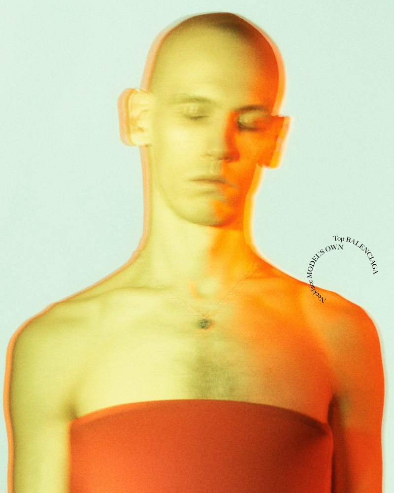 Portrait of a bald adult male with closed eyes and shoulder-length orange fabric around chest against a pale blue-gray studio backdrop.
