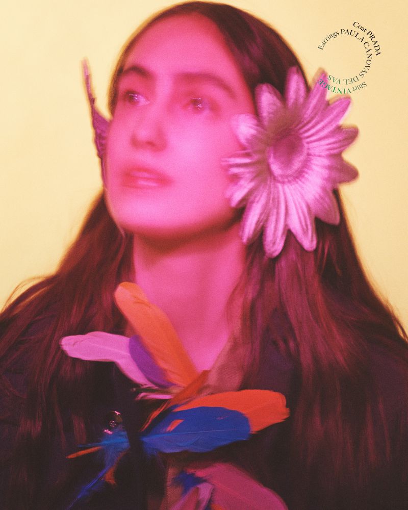 Portrait of a female model with a flower and feathers, wearing Prada earrings, with a yellow background and pink color grading.