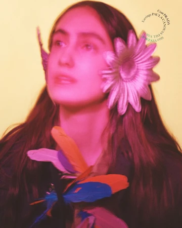 Portrait of a female model with a flower and feathers, wearing Prada earrings, with a yellow background and pink color grading.