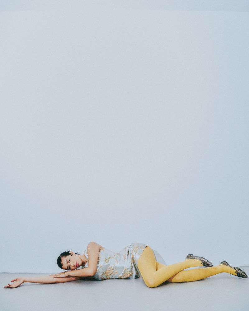 Woman lying on her side wearing a dress and yellow tights against a white background in a studio setting with neutral colour grading.