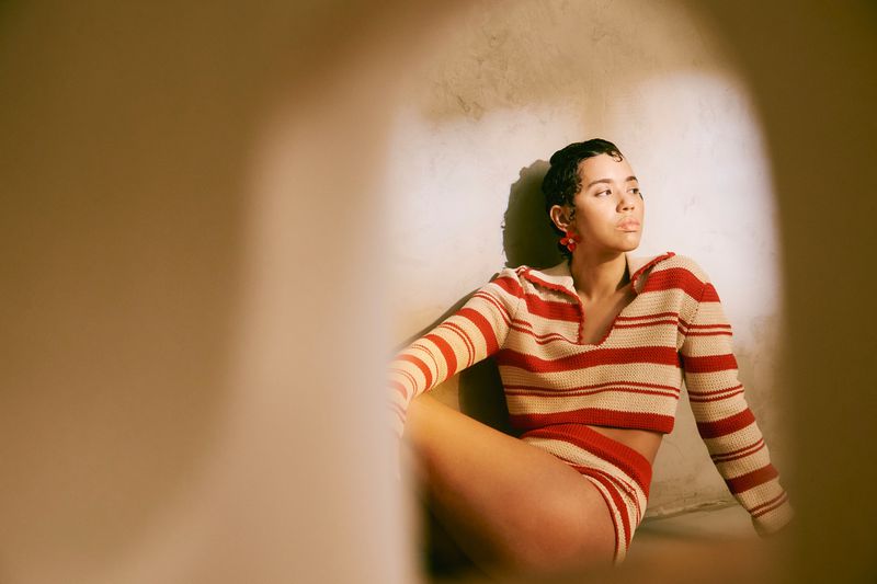 Jasmin Savoy Brown wearing a striped knit polo top and shorts against a neutral textured wall.