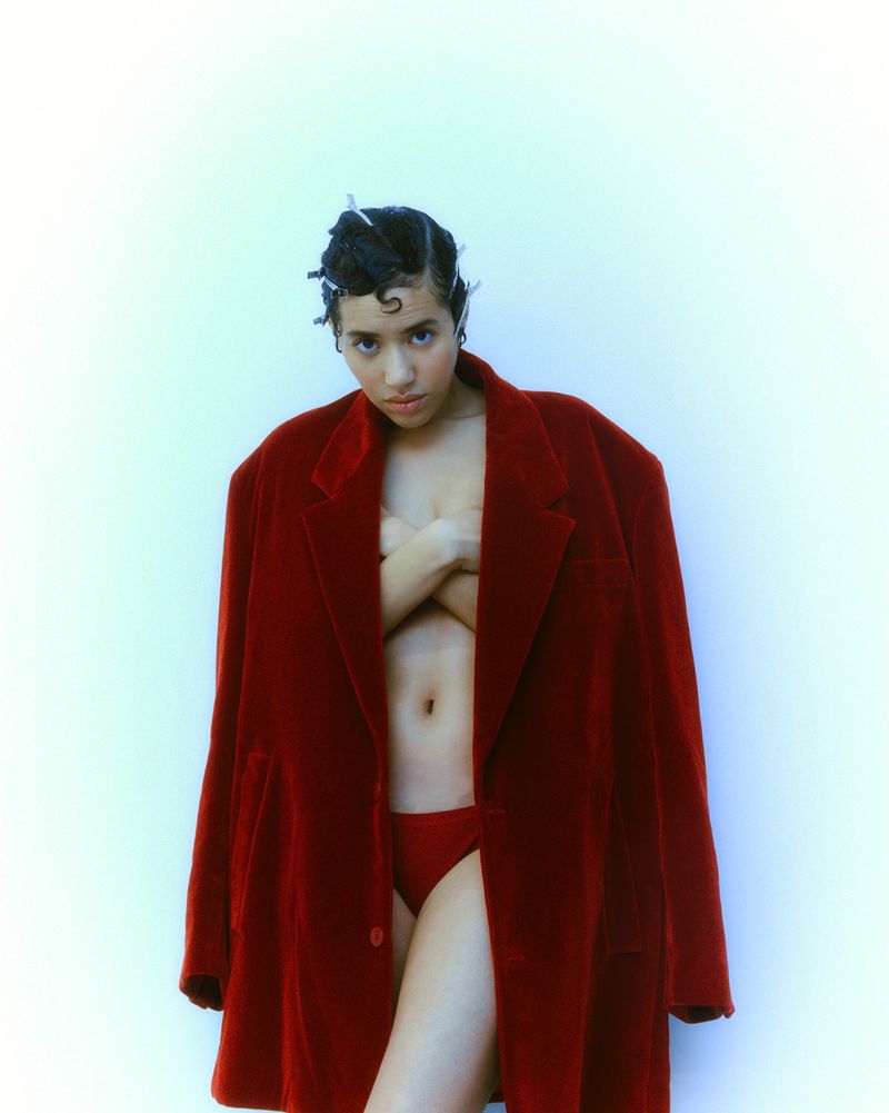 Young woman poses indoors wearing a large red blazer over a matching lingerie set, with a bright, soft-lit background.